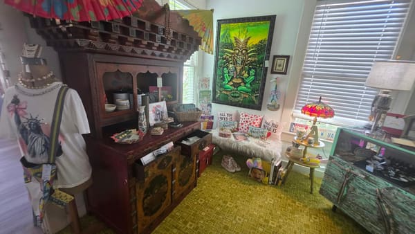 A carved wooden hutch with jewelry, books and "All You Need Is Love" pillows, beside a tiki painting and a stained-glass lamp