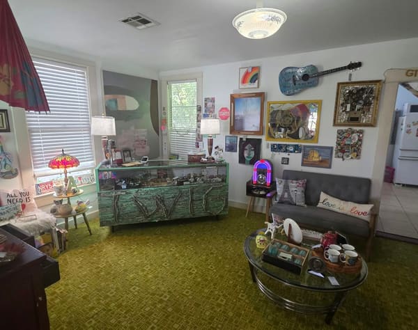A green-carpeted room with an art-covered wall, a painted guitar, a display case trimmed in twigs and a gray sofa