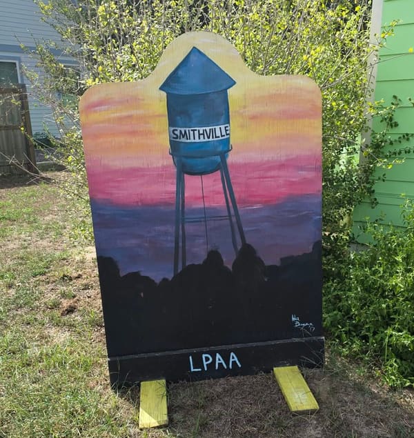 A hand-painted sign of the Smithville water tower against a sunset sky, signed LPAA, standing in the garden