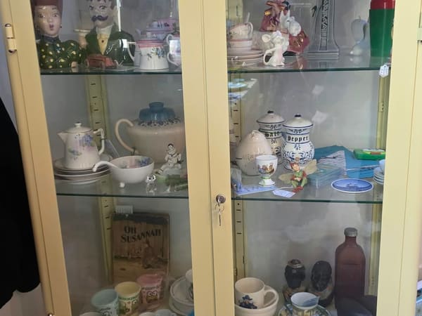 A pale yellow vintage curio cabinet filled with floral teapots, china cups and collectible figurines